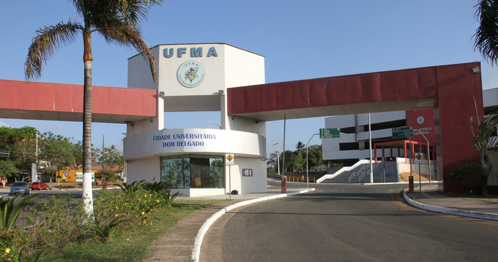 Fachada da UFMA com uma guarita de paredes brancas e vermelhas contendo a logo da universidade e o nome da cidade universitária Dom Delgado